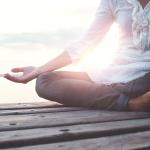Frau sitzt im Yogasitz am Steg am Wasser und macht Entspannungsübungen